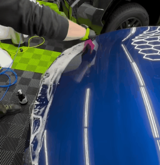 A worker uses a pink squeegee to smooth a protective film onto the shiny blue hood of a car in a detailing shop. The car’s surface reflects bright overhead lights, and the geometric design on the hood is partially visible. The shop’s green and black checkered floor can be seen in the background along with various detailing tools and equipment.