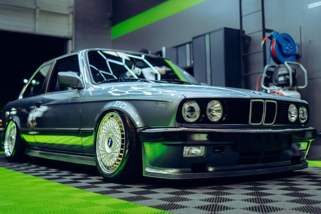 A classic gray BMW with detailed custom rims is parked inside a detailing shop. The car is low to the ground with a sleek, polished finish, reflecting the overhead lights. The car is set against a neon green and black checkered floor, and various tools and equipment can be seen on the walls in the background. The car’s front end, including its iconic grille and headlights, is prominently displayed.