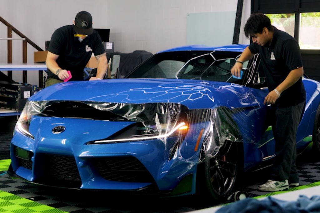 Two workers apply paint protection film (PPF) to the front of a blue sports car in a detailing shop. The film is being carefully smoothed over the hood and front bumper as one worker uses a pink squeegee tool and the other uses a blue tool. The car's hood features a geometric design, and the shop's interior is visible in the background with various equipment and tools.