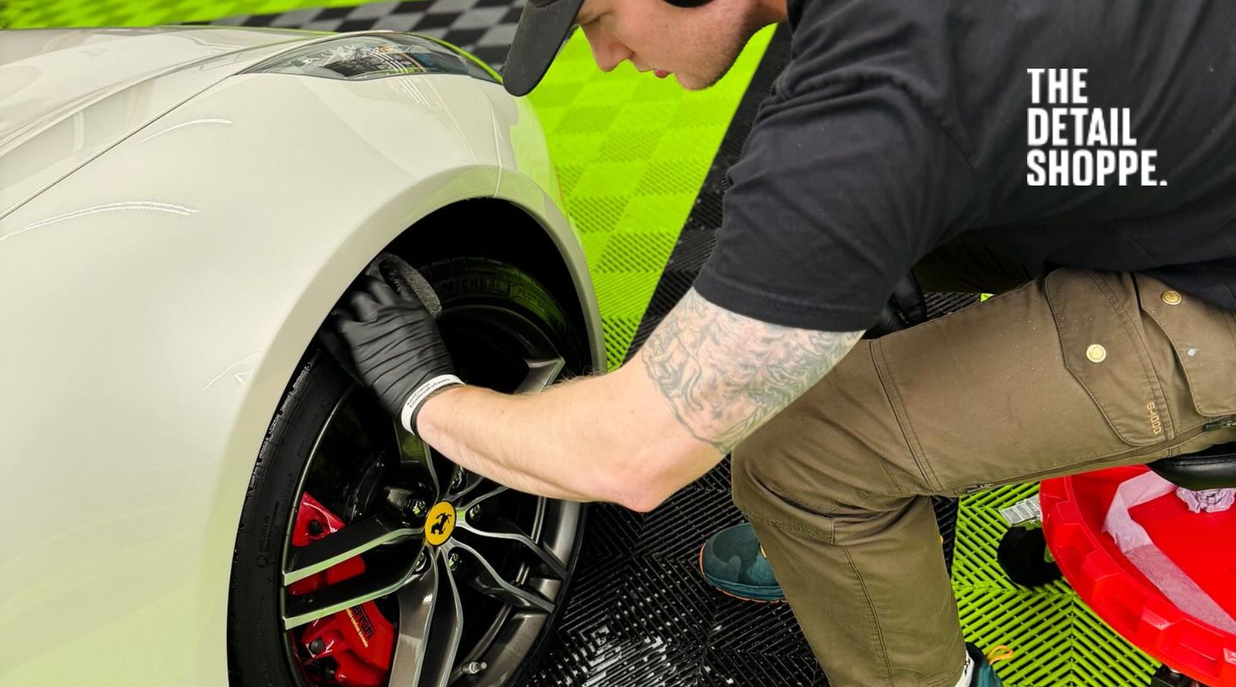 Best Exterior Detailing Services in Happy Valley - A man wearing gloves is polishing the wheel of a luxury car, which appears to be a Ferrari, with red brake calipers. The background features a checkered green and black floor mat. The logo "The Detail Shoppe" is displayed in the upper-right corner of the image.