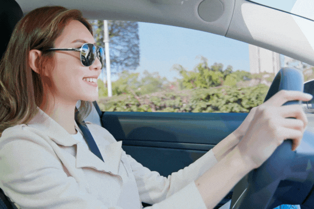 A woman wearing sunglasses smiles as she drives a car on a sunny day. The sunlight streams through the tinted side windows, indicating the use of UV protection tint for both safety and comfort.