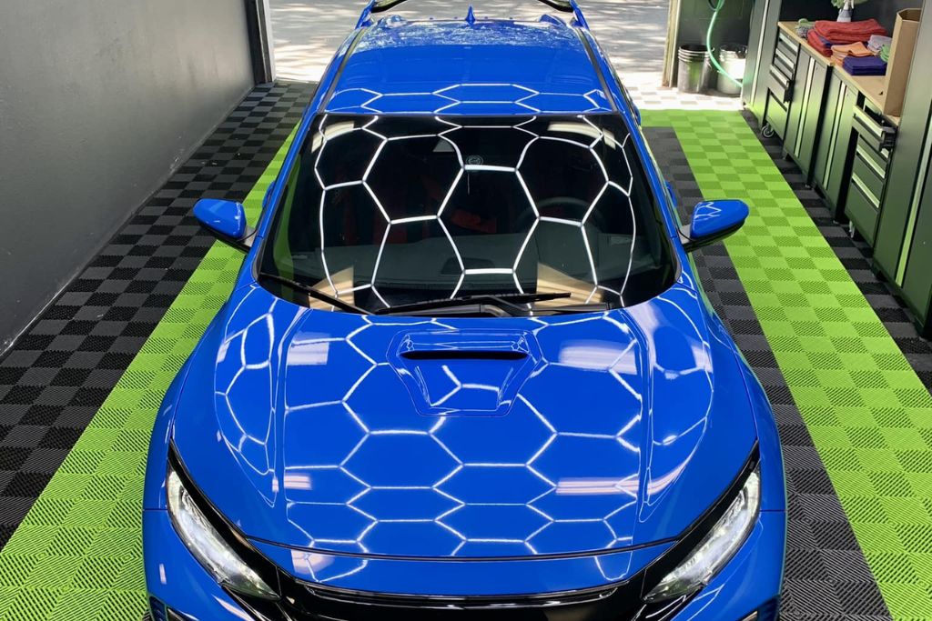 A vibrant blue car is parked in a detailing shop, with hexagonal overhead lights reflecting off its glossy surface. The car sits on a black and green checkered floor, with detailing equipment and towels visible on a workbench in the background. The vehicle's clean, polished finish showcases the results of professional detailing.