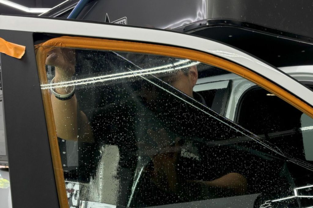 A person is carefully applying a tinted film to a car window, with water droplets visible on the glass. The tint installation process is in progress, highlighting the precision needed for proper UV and heat protection.