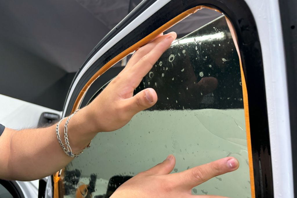 Close-up of hands carefully applying a tinted film to a car window. The window is partially covered in water droplets, indicating the tinting process is underway, with precision and care being applied.