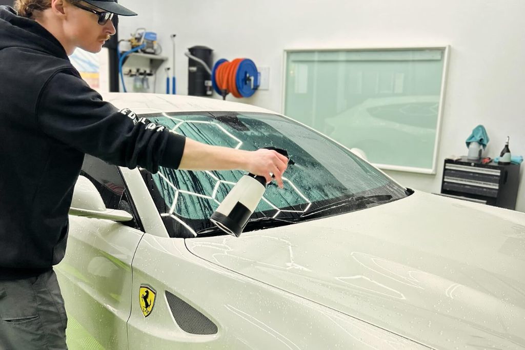 A person wearing sunglasses and a hoodie is spraying cleaning solution onto the windshield of a white Ferrari. The car is in a detailing shop, with cleaning equipment and supplies visible in the background, including coiled hoses and towels. The Ferrari's logo is clearly visible on the side of the car.