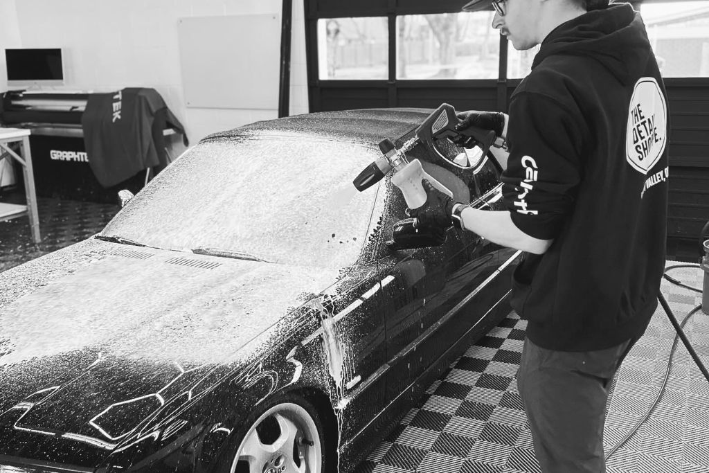 A person wearing a "The Detail Shoppe" hoodie and gloves is using a foam cannon to apply soap to a black car in a detailing shop. The car is covered in thick suds, and the shop features a checkered floor and equipment in the background. The scene captures the process of professional car washing and detailing.