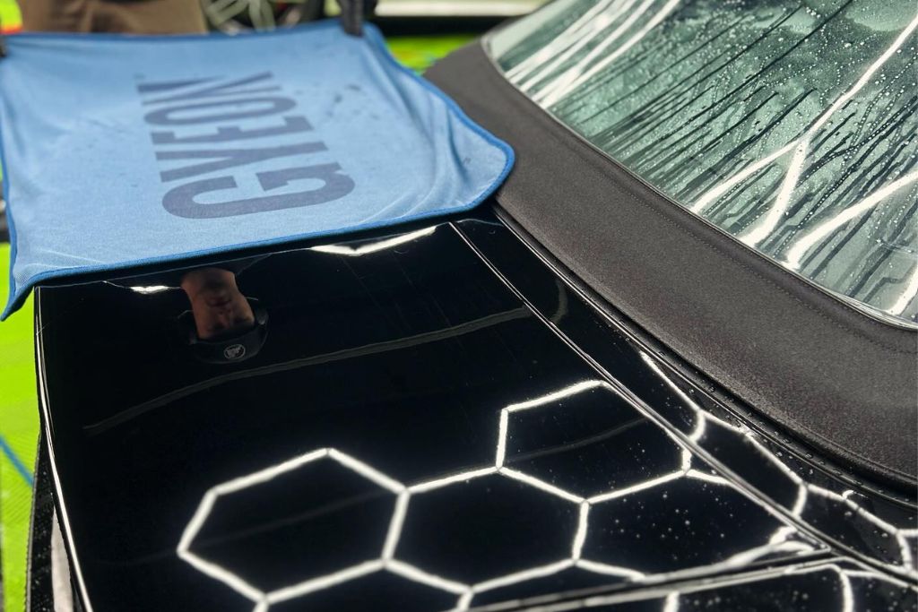 A black car with a polished surface reflects bright lighting, with a Gyeon towel placed on the trunk for detailing purposes. The car’s rear windshield shows droplets of water, emphasizing the importance of protective maintenance during winter.