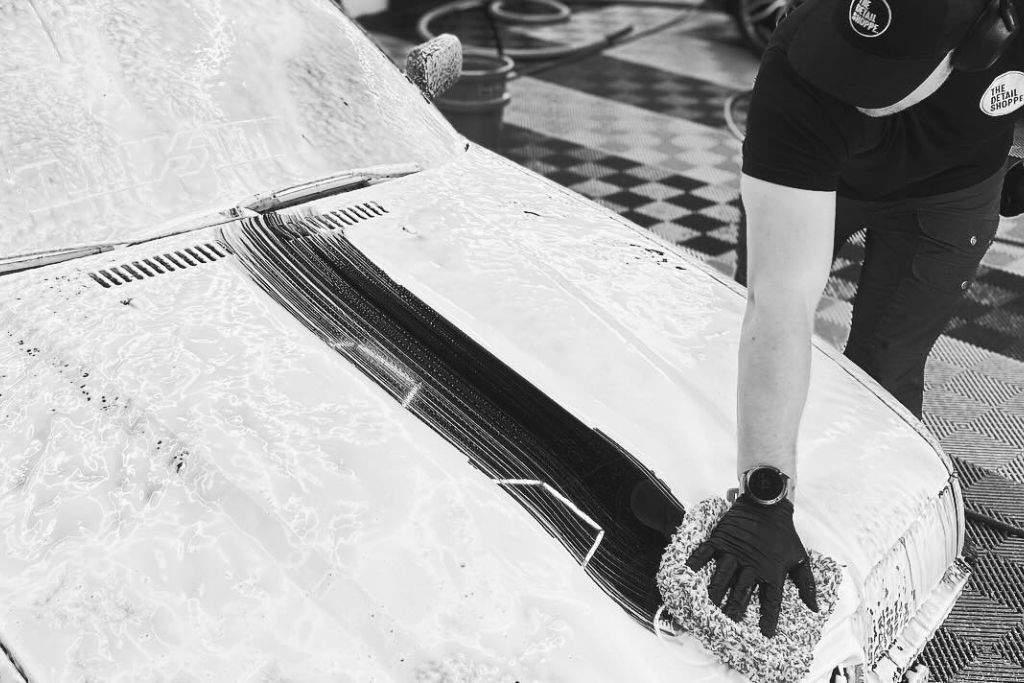 A person wearing gloves and a "The Detail Shoppe" cap carefully hand-washes the hood of a foam-covered car in a detailing shop. The setting features a clean, professional workspace with checkered flooring, emphasizing the attention to detail in car maintenance.