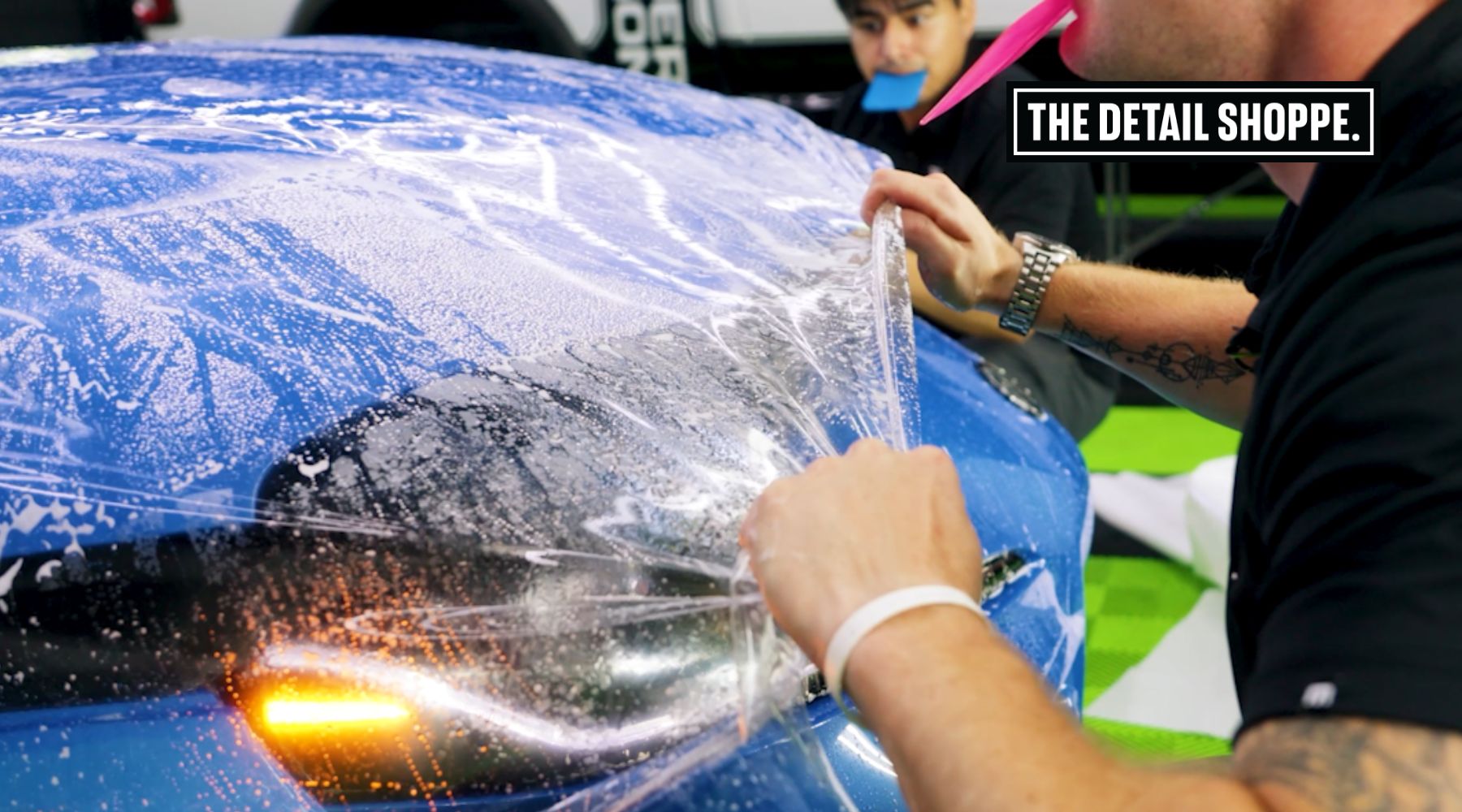 Close-up of a technician applying a clear paint protection film (PPF) onto a blue car hood covered in soapy water, with another technician in the background holding a tool in their mouth. The text "The Detail Shoppe" is visible in the upper corner.