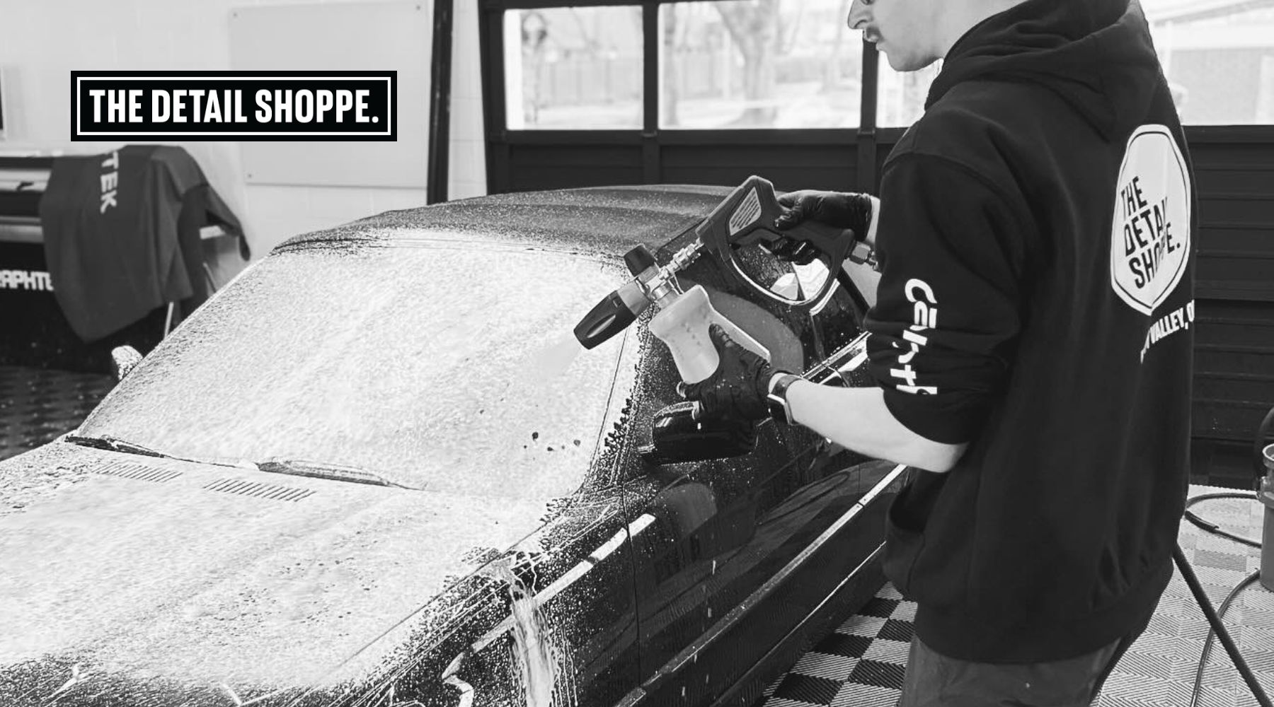 A person wearing a "The Detail Shoppe" hoodie sprays foam onto a black car using a detailing gun in a professional garage setting. The logo "The Detail Shoppe" is visible in the corner, emphasizing the business's branding.