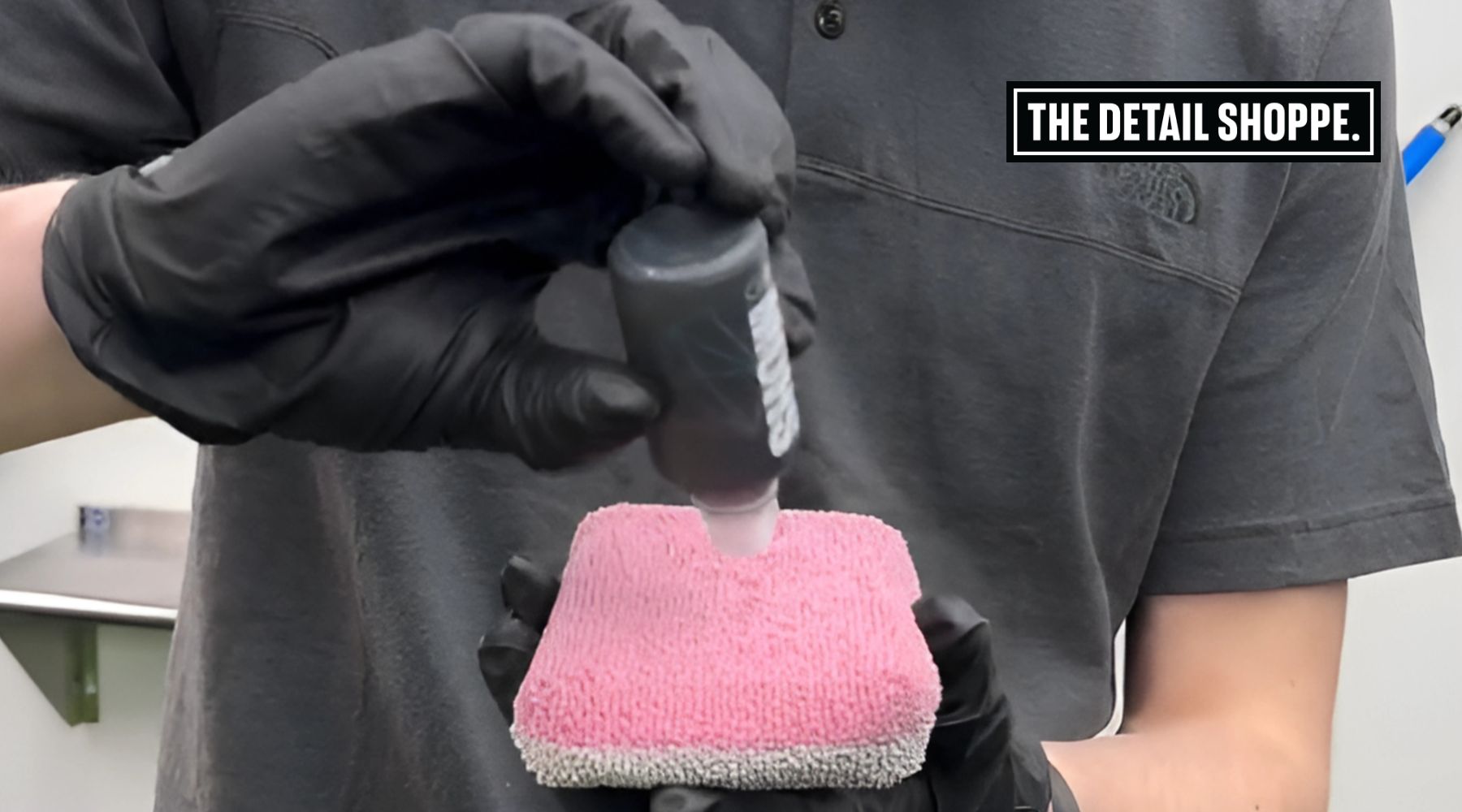 A person wearing black gloves applies ceramic coating liquid from a small bottle onto a pink microfiber applicator pad. The image is branded with a logo reading "The Detail Shoppe" in the top right corner.