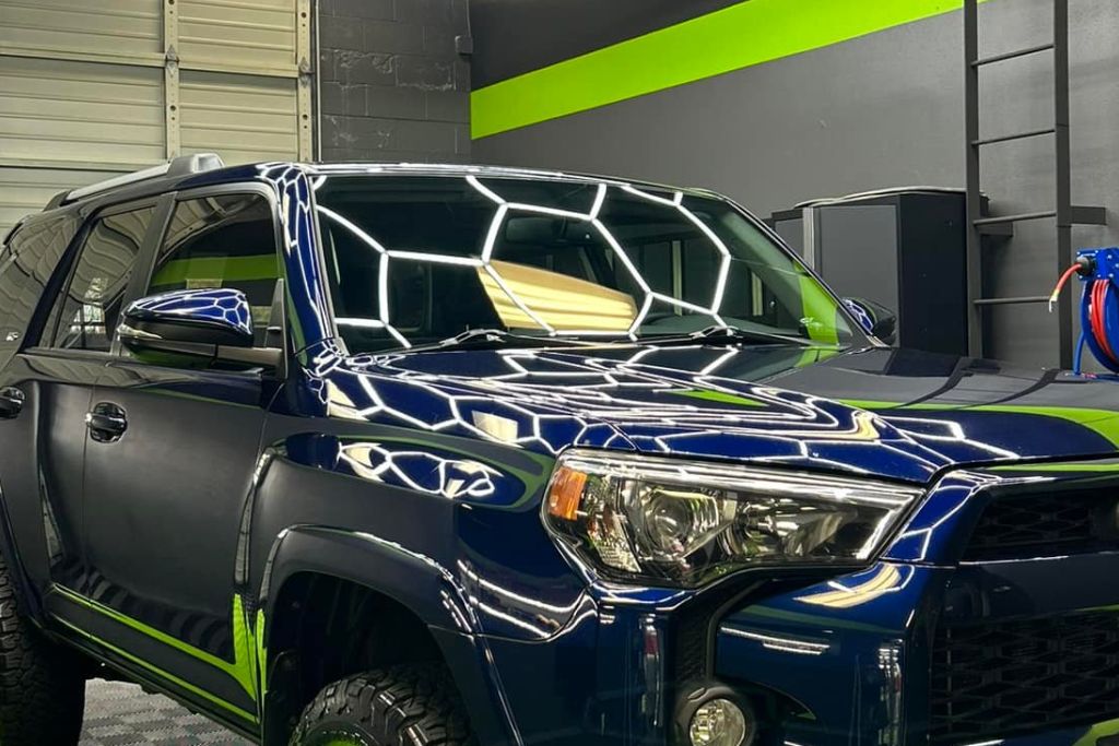 A shiny dark blue SUV with a mirror-like finish reflecting the hexagonal lighting in a detailing studio. The vehicle's flawless surface showcases the scratch-resistant properties of the ceramic coating.