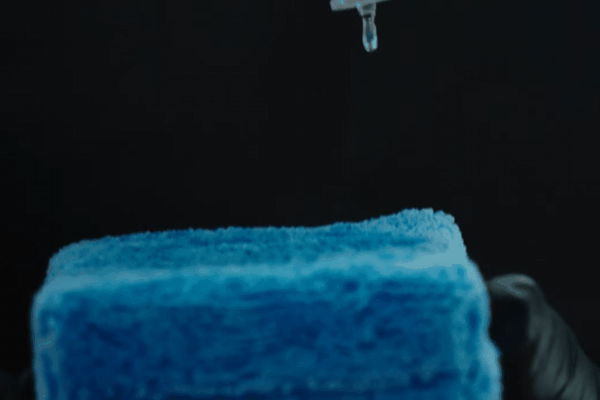 A close-up of a drop of ceramic coating liquid falling onto a blue microfiber applicator pad, held by a gloved hand against a dark background. The liquid is shown mid-drip, highlighting the precision of the application process.