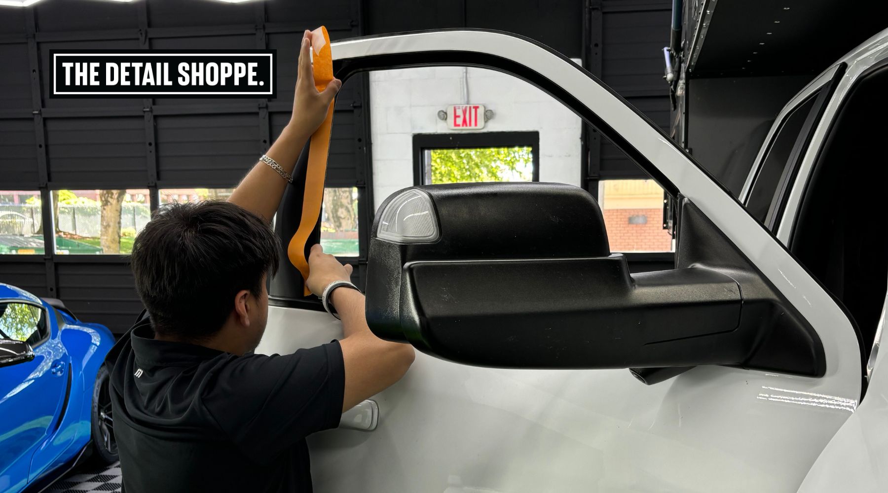 A detailing professional applies masking tape to the window frame of a white truck inside a garage. The workspace has a black interior with large windows, a visible "EXIT" sign, and a blue sports car parked in the background. A bold sign reading "THE DETAIL SHOPPE." is displayed on the wall.