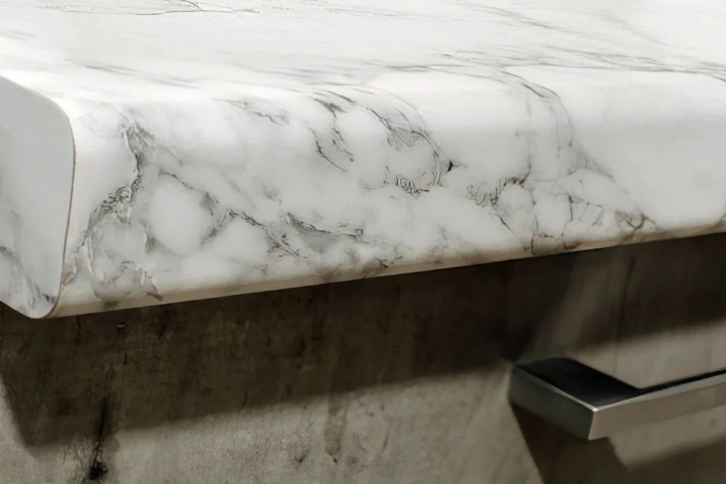 A close-up view of a quartz or laminate countertop with a rounded edge featuring a white and gray marble-like design. The smooth surface shows fine detailing, with a modern drawer handle visible below.