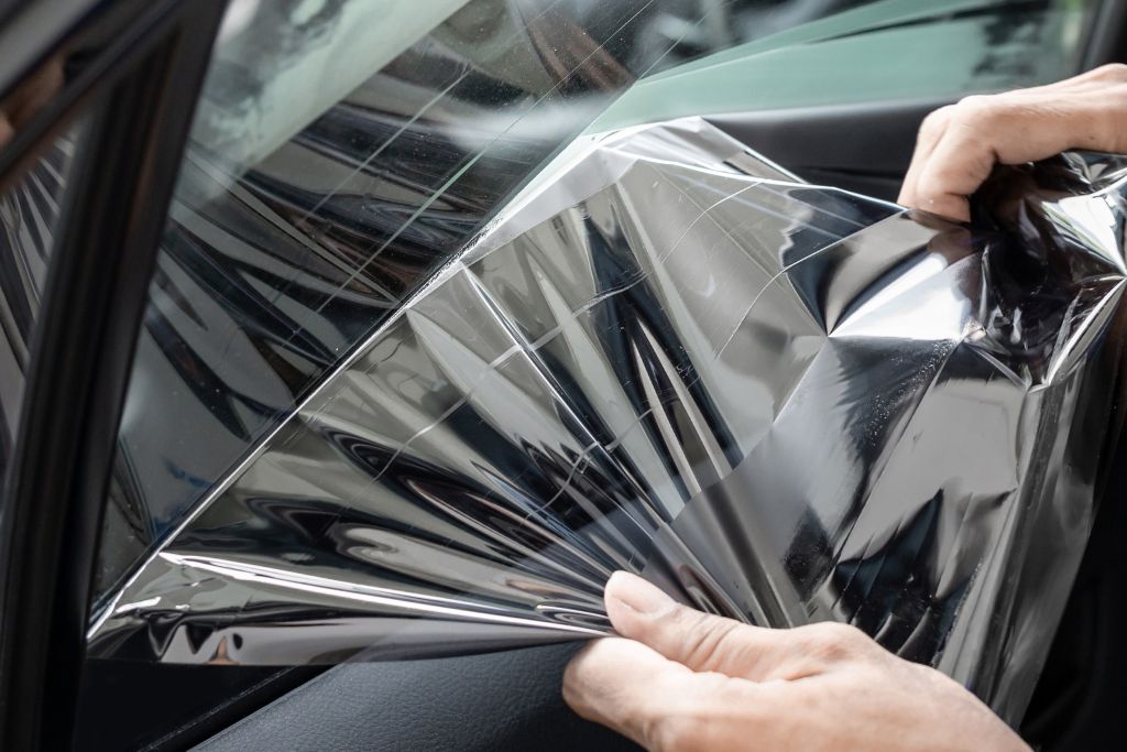 A close-up of hands peeling off or installing a tinted window film on a car door. The reflective film provides UV protection, heat rejection, and enhanced privacy. Window tinting options vary based on performance, aesthetics, and security needs.