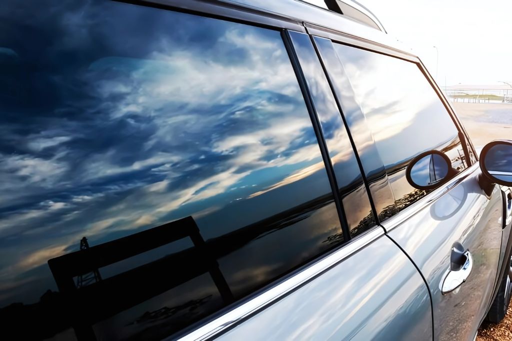 A close-up of a car with dark tinted windows reflecting a dramatic sky with clouds and sunlight. The deep tint provides privacy and security by obscuring the view inside the vehicle. The sleek design and reflective glass enhance both aesthetics and protection.