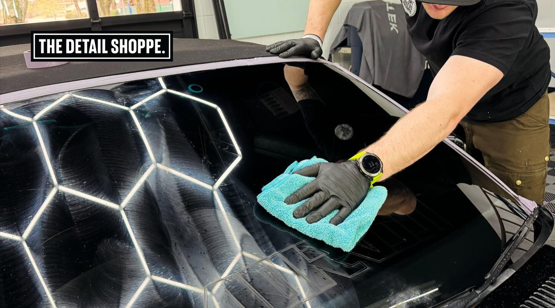 A professional detailer wearing black gloves and a smartwatch uses a microfiber towel to clean and polish a car windshield. The glass reflects hexagonal overhead lights, and the workspace appears to be a detailing shop. A "The Detail Shoppe" sign is prominently displayed in the upper left corner.