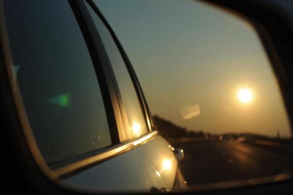 A car's side mirror reflects the sunset on an open road, with the sun casting a golden glow on the tinted windows. The smooth highway stretches into the distance, emphasizing the warmth and intensity of the sunlight. The tinted glass subtly reduces glare, highlighting the benefits of UV protection for both the driver and the vehicle.