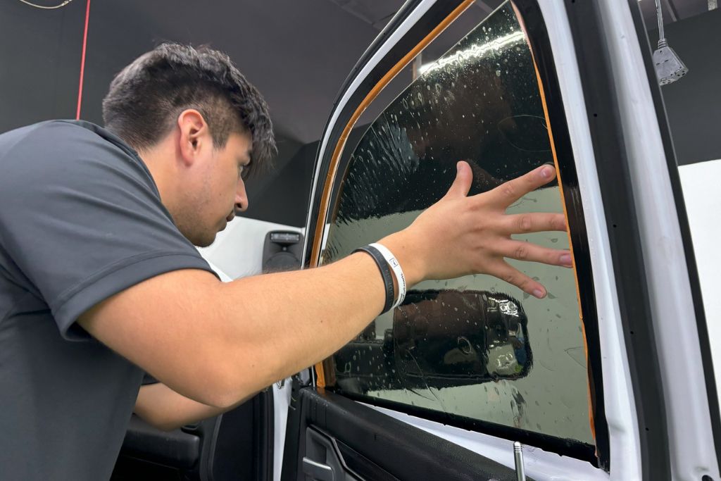 A professional installer carefully applies a tinted film to a car window, smoothing it out with his hand. The window is wet, indicating the preparation process for a seamless application. His focused expression and attention to detail highlight the precision required for high-quality tint installation.