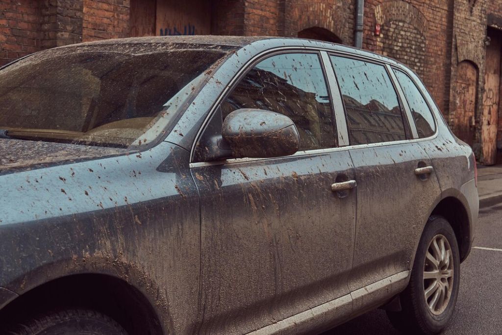 A dark-colored SUV is covered in mud and dirt, parked on a wet street against the backdrop of an old brick building. Mud splatters are visible on the windows, side mirrors, and body, emphasizing the impact of rough road conditions. The image highlights the need for protective solutions like Paint Protection Film (PPF) to maintain a vehicle’s appearance.