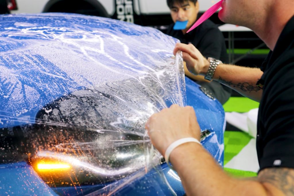 A professional detailer carefully applies Paint Protection Film (PPF) to the headlight and hood of a blue sports car. The transparent film is stretched and smoothed over the wet surface, ensuring a precise, bubble-free installation. Another technician in the background holds a squeegee in their mouth, preparing to assist with the application.