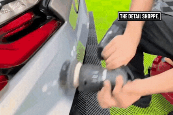 A detailing technician at The Detail Shoppe uses a dual-action polisher on the rear bumper of a silver car, prepping the surface for ceramic coating on a vibrant green and black checkered floor.