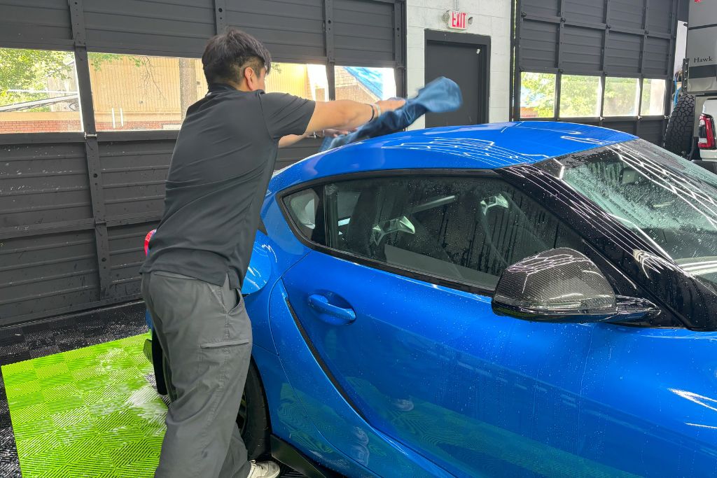 A detailing technician gently pats down a bright blue sports car with a microfiber towel, drying it after a wash while checking the surface and edges for signs of PPF lifting or damage.