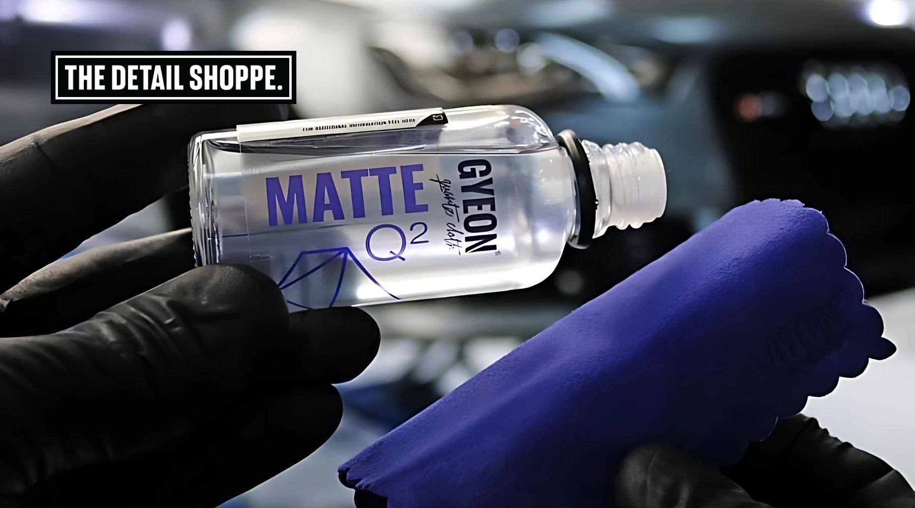 A gloved hand holds a bottle of Gyeon Q² Matte ceramic coating above a blue applicator pad, with the words "THE DETAIL SHOPPE." displayed in the background. The bottle is labeled for matte finishes, indicating it's tailored for protecting non-glossy vehicle surfaces.