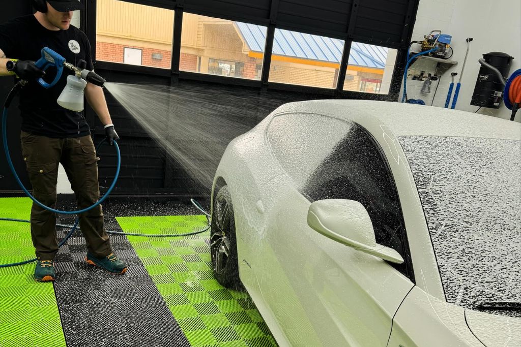 A detailing technician at The Detail Shoppe hand-sprays thick foam soap onto a white sports car during a PPF-safe wash. The vehicle is covered in suds, demonstrating proper maintenance techniques for preserving paint protection film.