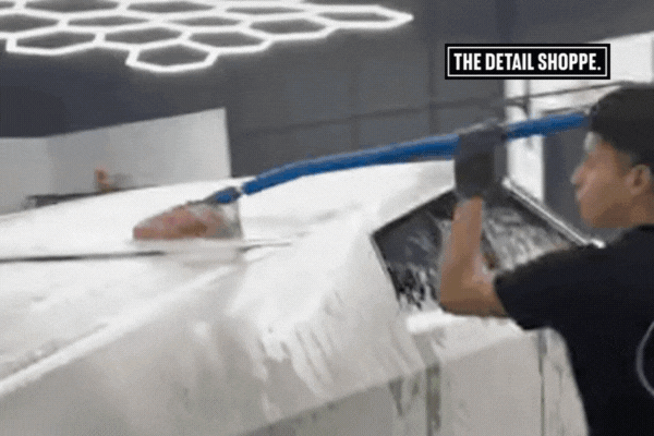 A technician from The Detail Shoppe professionally hand-washes a foam-covered white vehicle under clean, modern lighting—demonstrating expert care and PPF-safe techniques.
