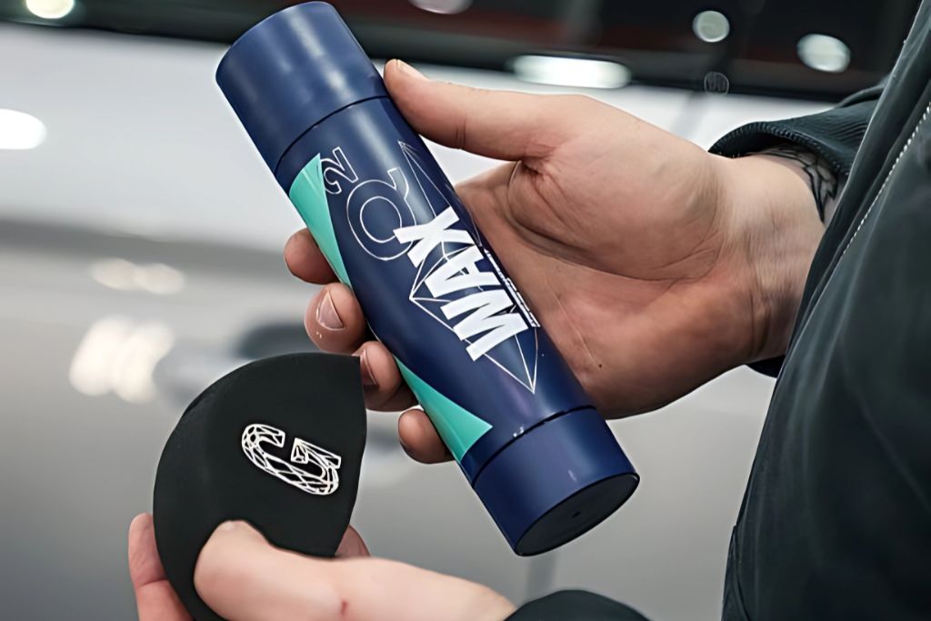 Close-up of a person holding a dark blue bottle labeled "WAX" and a black applicator pad with a stylized “G” logo, preparing for automotive waxing. The background shows a glossy car surface, suggesting the context of paint or PPF maintenance.