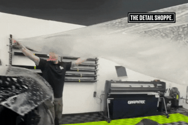 A detailing professional stretches a large sheet of clear paint protection film (PPF) high above a vehicle, preparing it for installation in a clean, high-tech workspace at The Detail Shoppe.