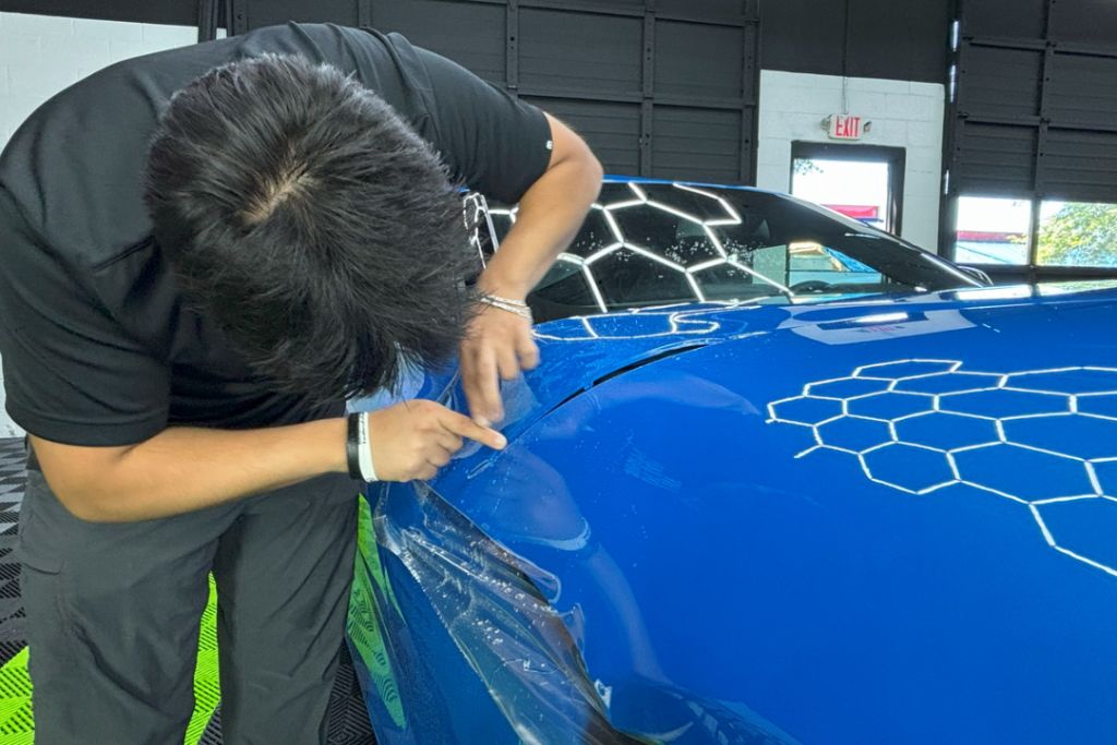 Close-up of a technician using a precision tool to tuck and seal paint protection film (PPF) along the edge of a blue sports car’s front fender, highlighting the detail and skill involved in quality installation.