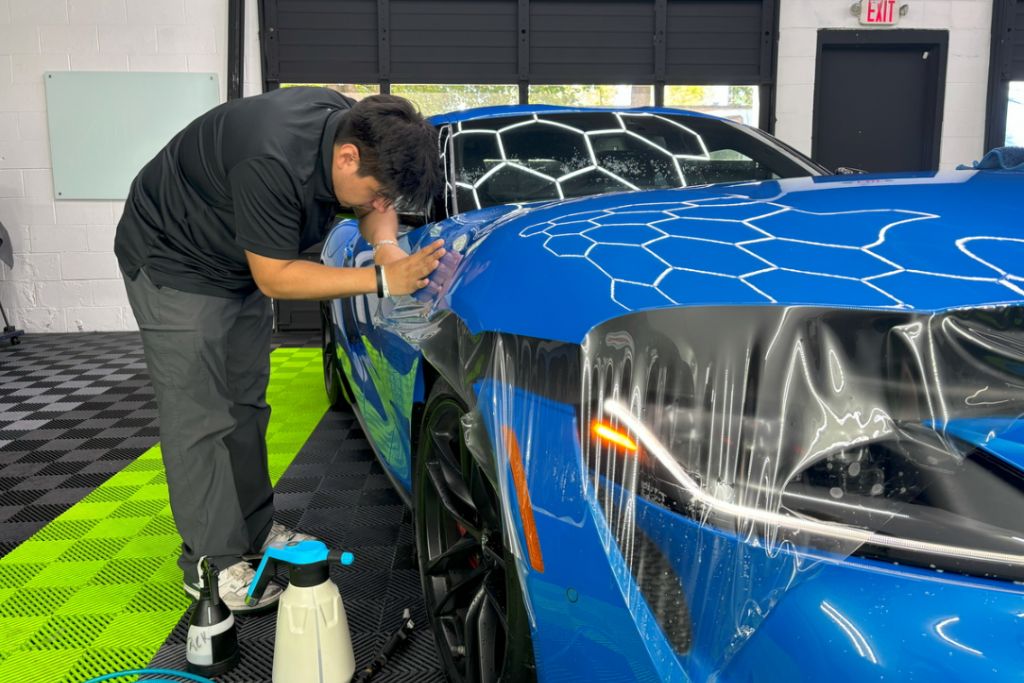 A detailing technician carefully applies paint protection film (PPF) to the front fender of a bright blue sports car inside a modern garage, with portions of the clear film visibly stretched and smoothed over the paint.