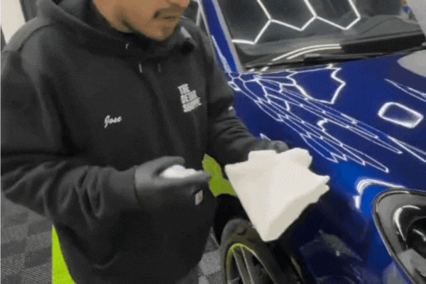 Detailing technician at The Detail Shoppe wearing gloves and holding a spray bottle and microfiber towel, preparing to wipe down a vibrant blue vehicle under LED lights. The image illustrates the hands-on process involved in understanding ceramic coating removal.