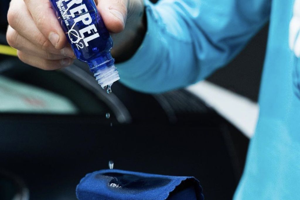 A close-up of a person in a blue shirt applying a few drops of ceramic coating from a blue "REPEL" bottle onto an applicator pad. This shows the precise process involved in maximizing the durability of ceramic coating on a new vehicle.