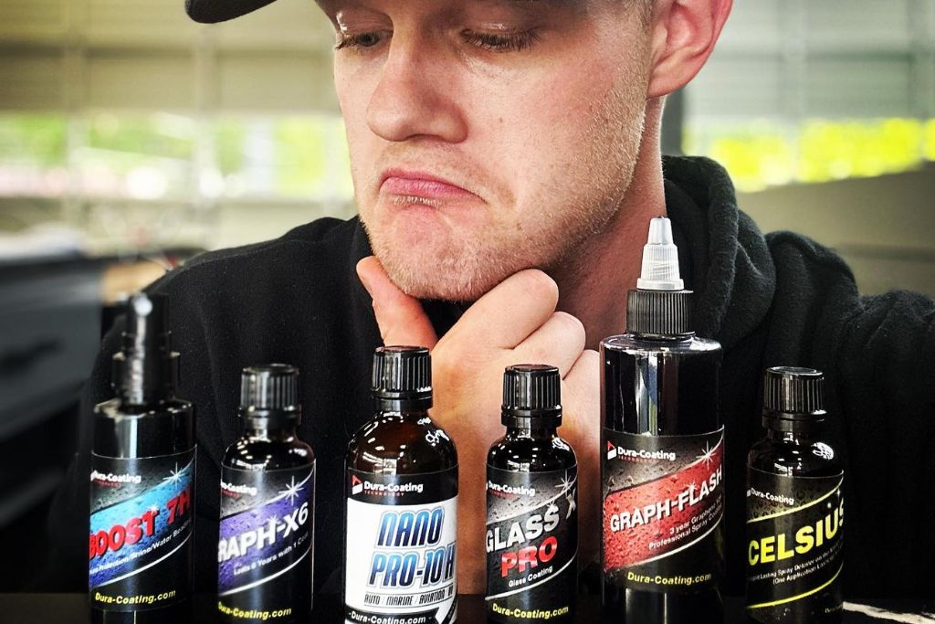 A man in a black hoodie and cap looks thoughtfully at six different Dura-Coating ceramic coating bottles lined up in front of him, reflecting on various product options to determine if ceramic coating is worth it.