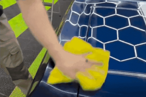 A person uses a bright yellow microfiber cloth to polish a car hood coated with ceramic, revealing a glossy blue finish with hexagonal reflections. This highlights the sleek, protective shine created by ceramic coating for new cars.