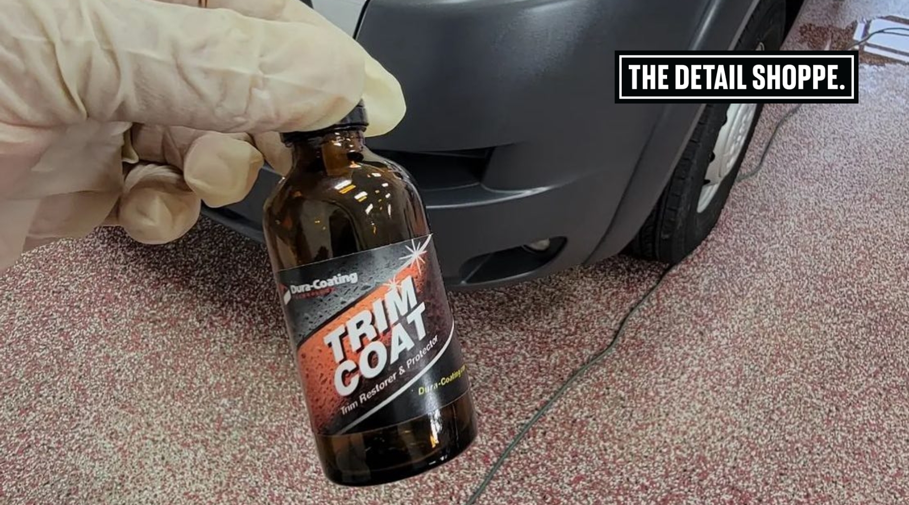 Gloved hand holding a bottle of Dura-Coating Trim Coat in front of a car bumper at The Detail Shoppe in Happy Valley, Oregon, showing ceramic coating prep during fall maintenance.