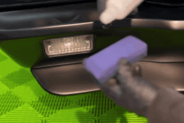A gloved hand applies ceramic coating to a vehicle's bumper using a purple applicator pad while liquid is dispensed from a white bottle. This close-up shows the first step in the curing process, illustrating why ceramic coating needs time to cure for full bonding and protection, especially in humid climates like Happy Valley, OR.