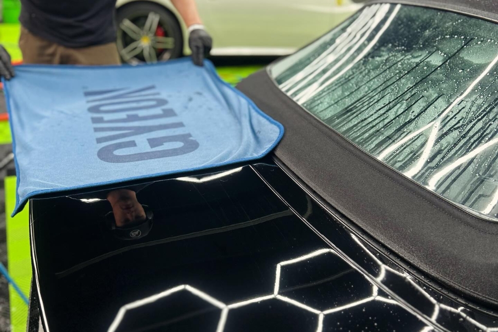A detailing technician gently lays a blue GYEON microfiber towel over the rear of a freshly ceramic-coated black convertible inside a garage. The image demonstrates how to protect your vehicle from rain after ceramic coating by blotting, not wiping, to preserve the finish during curing—crucial in damp climates like Happy Valley, OR.