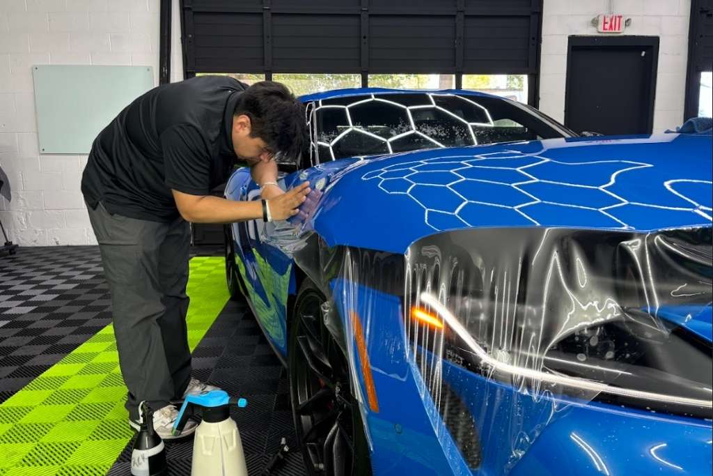 A technician at The Detail Shoppe in Happy Valley, OR carefully smooths paint protection film over the front fender of a vibrant blue sports car. This image illustrates how professional installation plays a key role in how long Paint Protection Film Typically Lasts, especially in climates like Oregon’s.