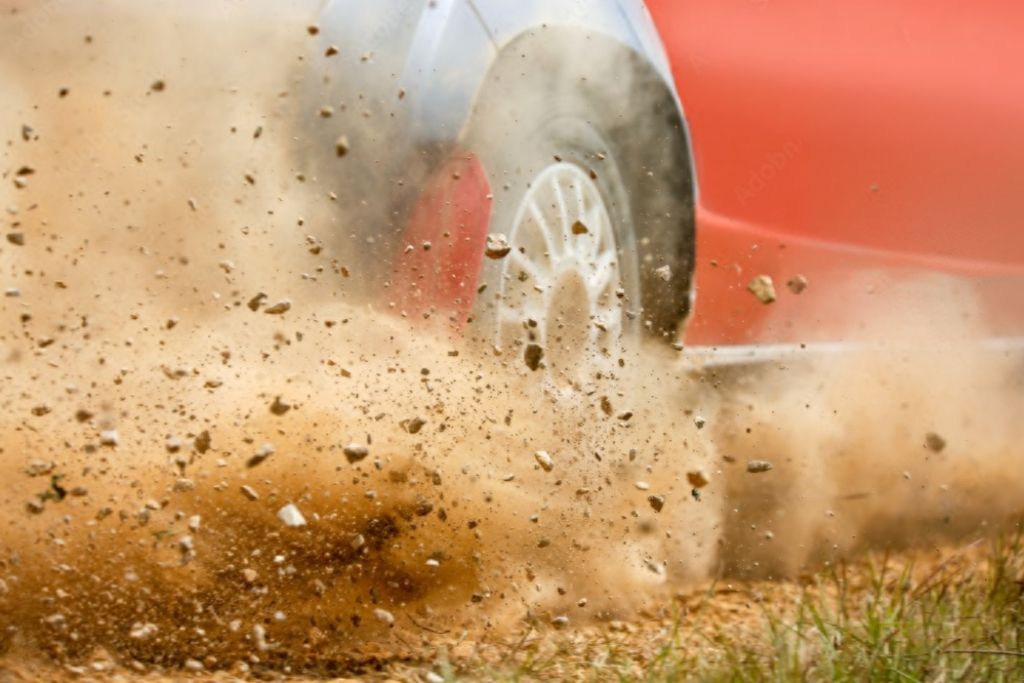 Close-up of a red vehicle's rear tire kicking up dirt, gravel, and small rocks while driving off-road, illustrating the kind of debris that Paint Protection Film helps shield against.