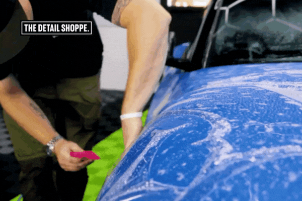 An installer at The Detail Shoppe in Happy Valley, OR uses a pink squeegee to smooth out paint protection film on the fender of a blue car covered in application solution. The motion and detail showcase The Detail Shoppe's Expertise in delivering flawless, professional PPF application with precision and care.