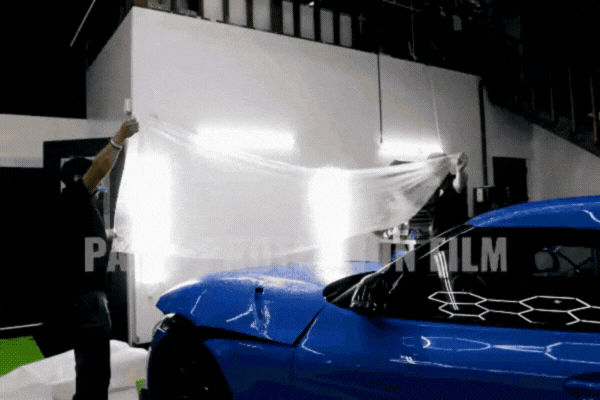 A technician at The Detail Shoppe in Happy Valley, OR carefully stretches a large sheet of clear paint protection film over the hood of a blue sports car under bright workshop lights. This image illustrates the Basics of PPF as a transparent barrier that protects high-impact areas of a vehicle from damage.