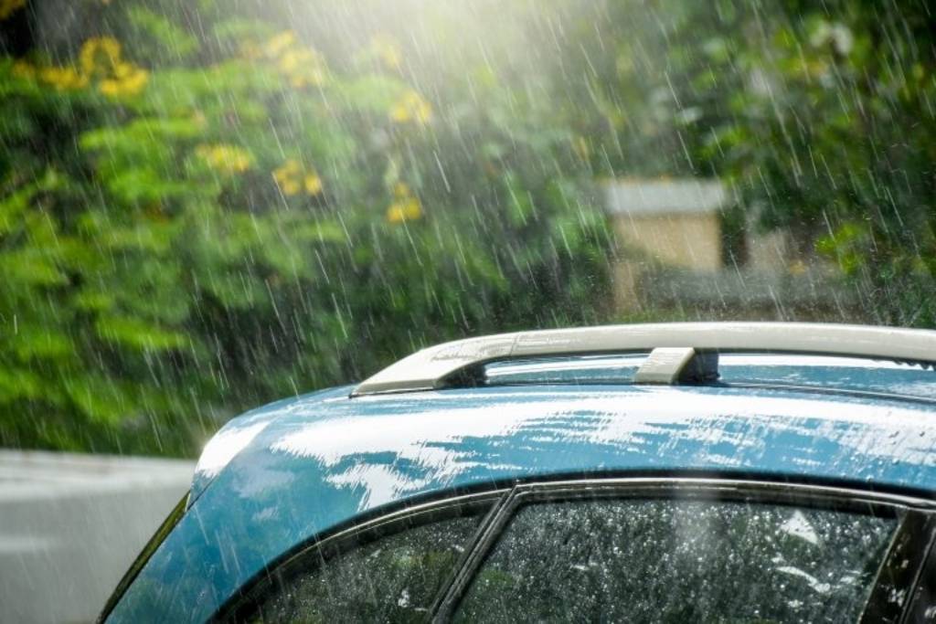 Close-up of rain pouring over the roof and windows of a parked blue SUV surrounded by greenery in Oregon. This visual represents how year-round moisture and weather conditions in the Pacific Northwest affect PPF lifespan, particularly when proper installation and film quality are lacking.
