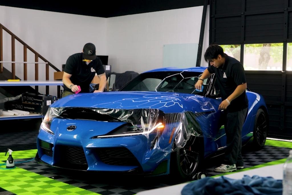 Two technicians at The Detail Shoppe in Happy Valley OR apply Paint Protection Film to a blue Toyota Supra using precision tools and techniques to ensure a smooth installation on the hood and front fenders.