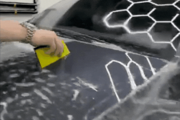 Close-up of a technician at The Detail Shoppe in Happy Valley, OR, using a yellow squeegee to smooth Paint Protection Film onto a black car hood under hexagon-patterned studio lights, demonstrating the precise PPF installation process.