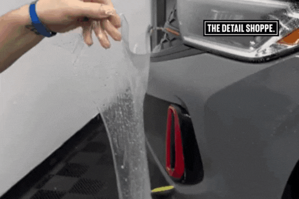 A technician at The Detail Shoppe in Happy Valley OR holds a clear sheet of Paint Protection Film, preparing to apply it to the front bumper of a gray vehicle with precision and control.
